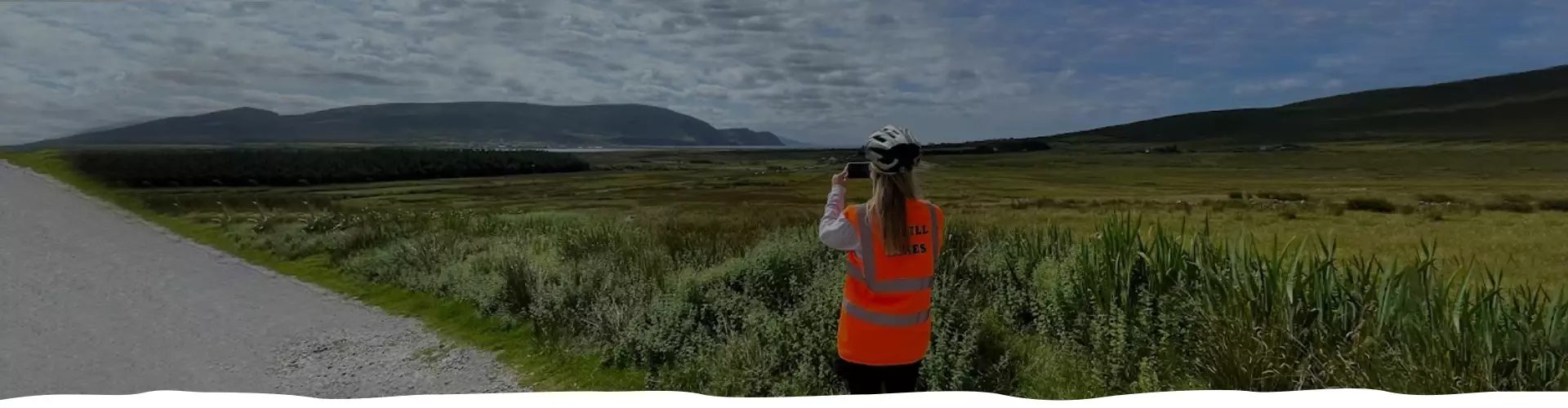 Person in orange vest using binoculars by the roadside with expansive grassy fields and hills in background.