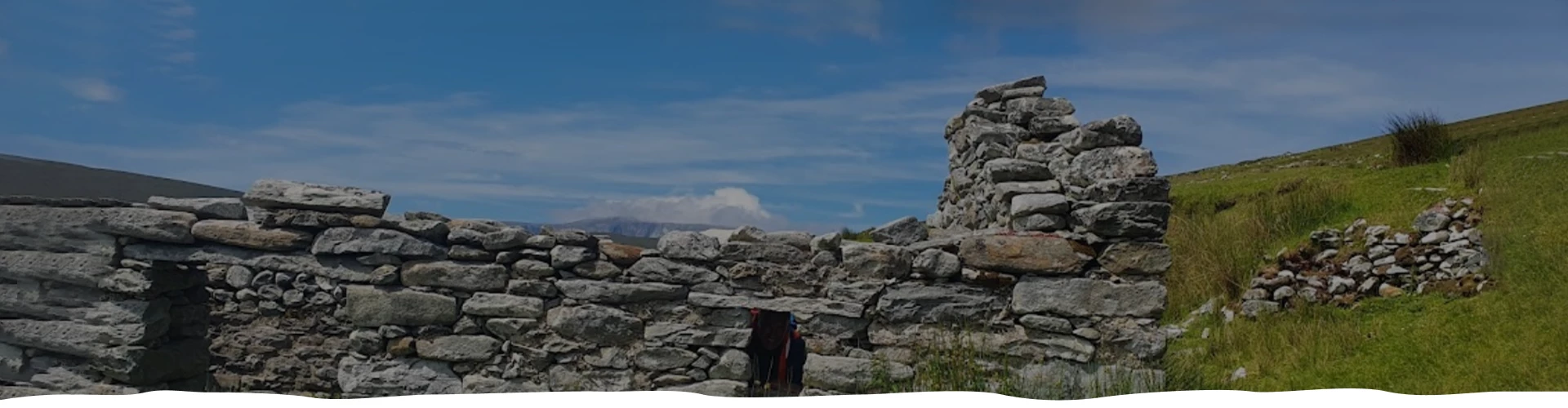 Ruins of an ancient stone structure under a clear blue sky, surrounded by grassy fields.