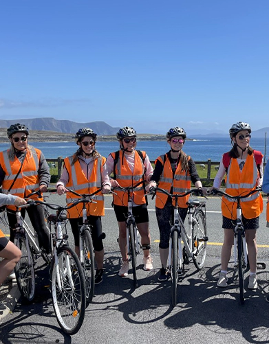 Group of five cyclists in orange vests standing with their bikes on a sunny coastal road.