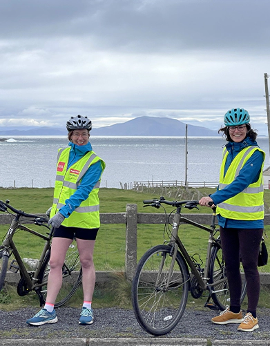 Two cyclists in reflective vests stand with bikes by a coastal area with a mountain in the background.