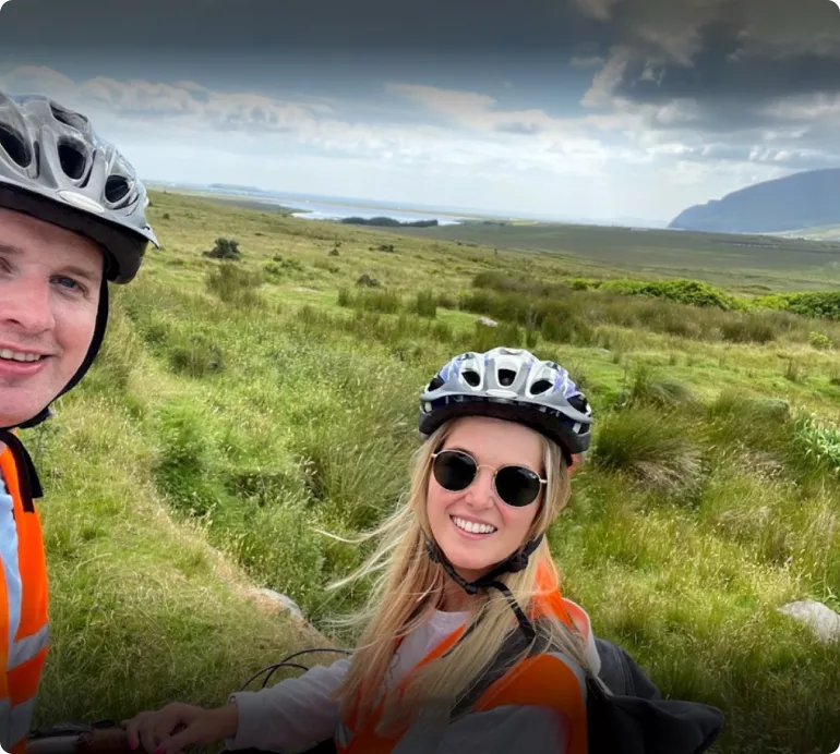Two cyclists wearing helmets and high-visibility vests smiling in a grassy field under a cloudy sky.