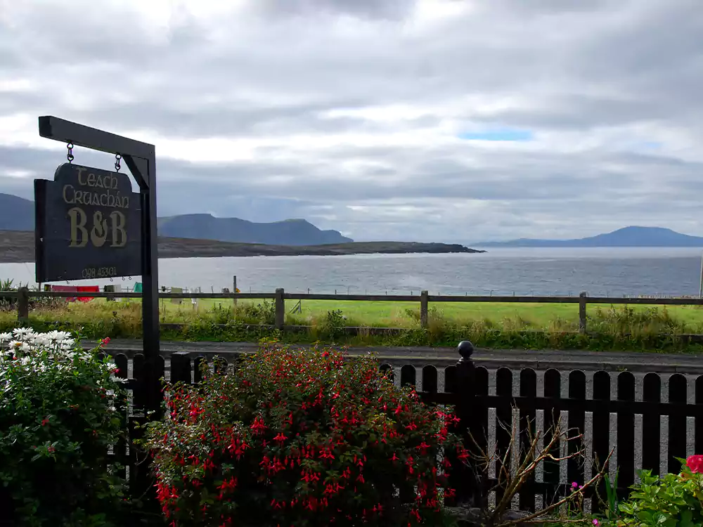 Seaside B&B sign with ocean and mountains in the background, surrounded by a flowered fence.
