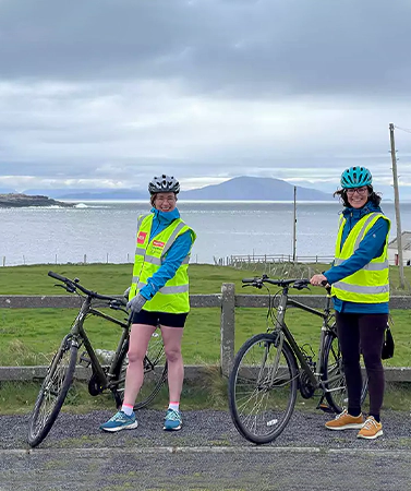 Two cyclists in reflective vests standing with bikes, coastal landscape with mountains in the background.