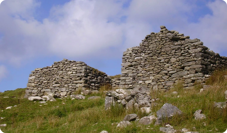 Ruined stone structures on a grassy hillside under a blue sky with clouds.