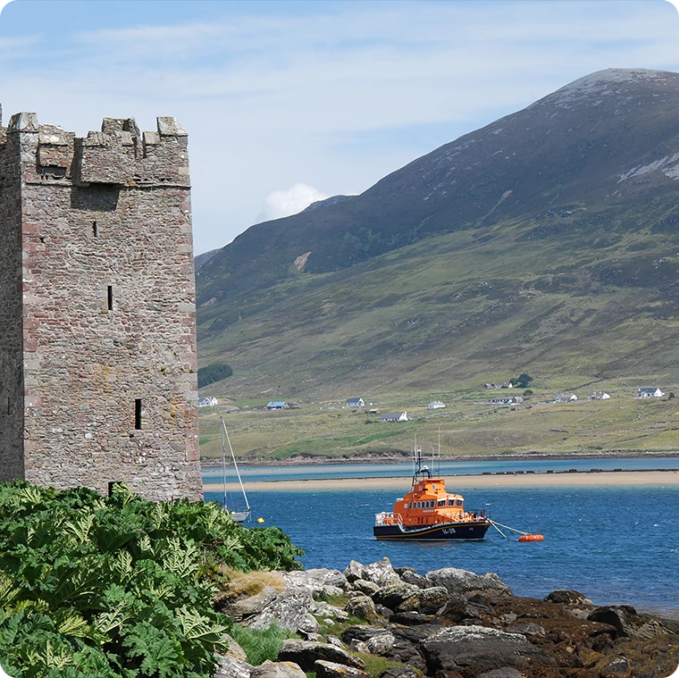 Ancient stone tower overlooking a bay with a lifeboat, surrounded by lush green hills.