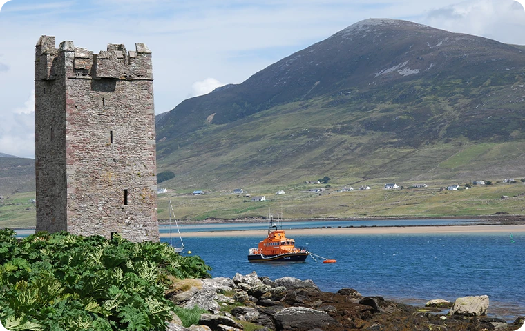 An ancient tower stands by a bay with a rescue boat and mountain background.